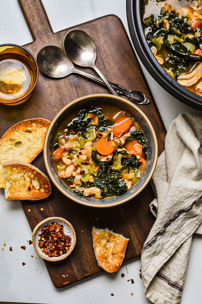 Bowl of chicken bean and kale soup with bread next to it.