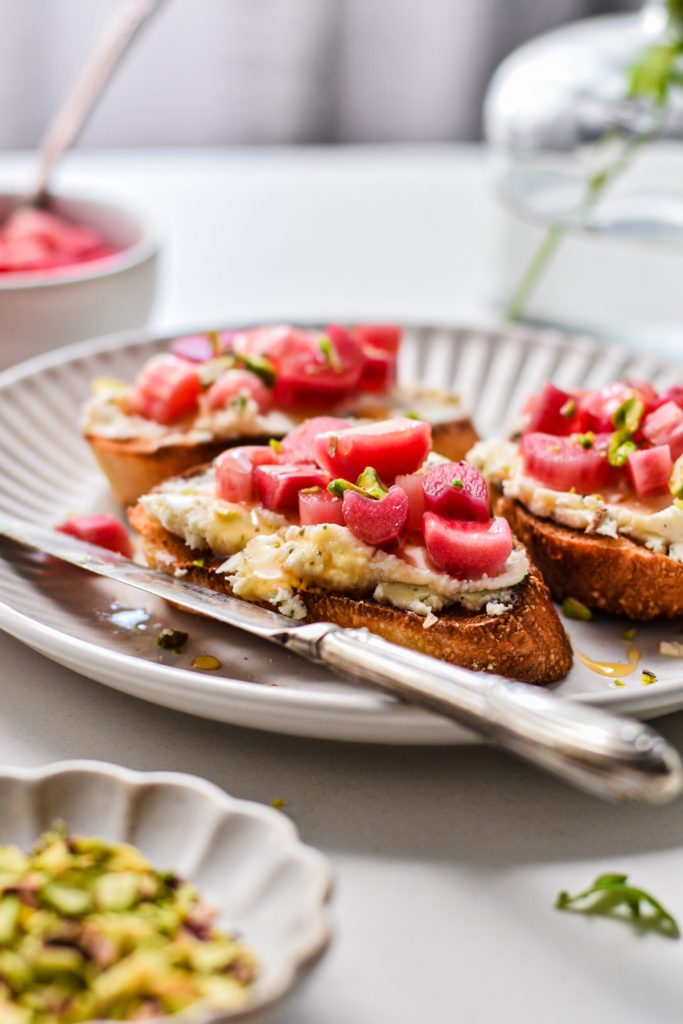 Side view of toasted pieces of bread topped with cheese, pistachios, pickled rhubarb and honey.