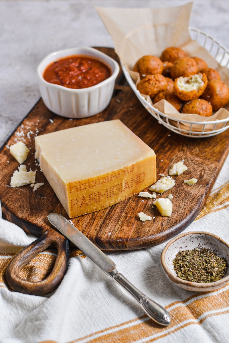 Block of parmesan cheese on a wooden board with the polpette di parmigiano behind it.