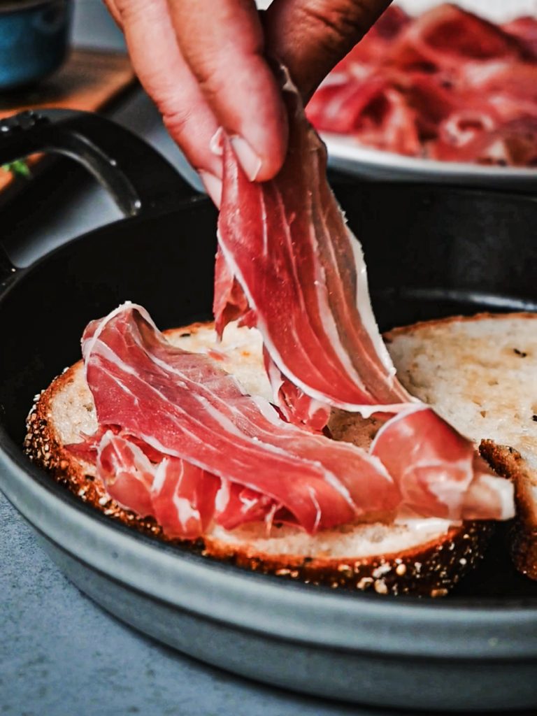 Hand adding prosciutto on top of the pieces of bread in a skillet.