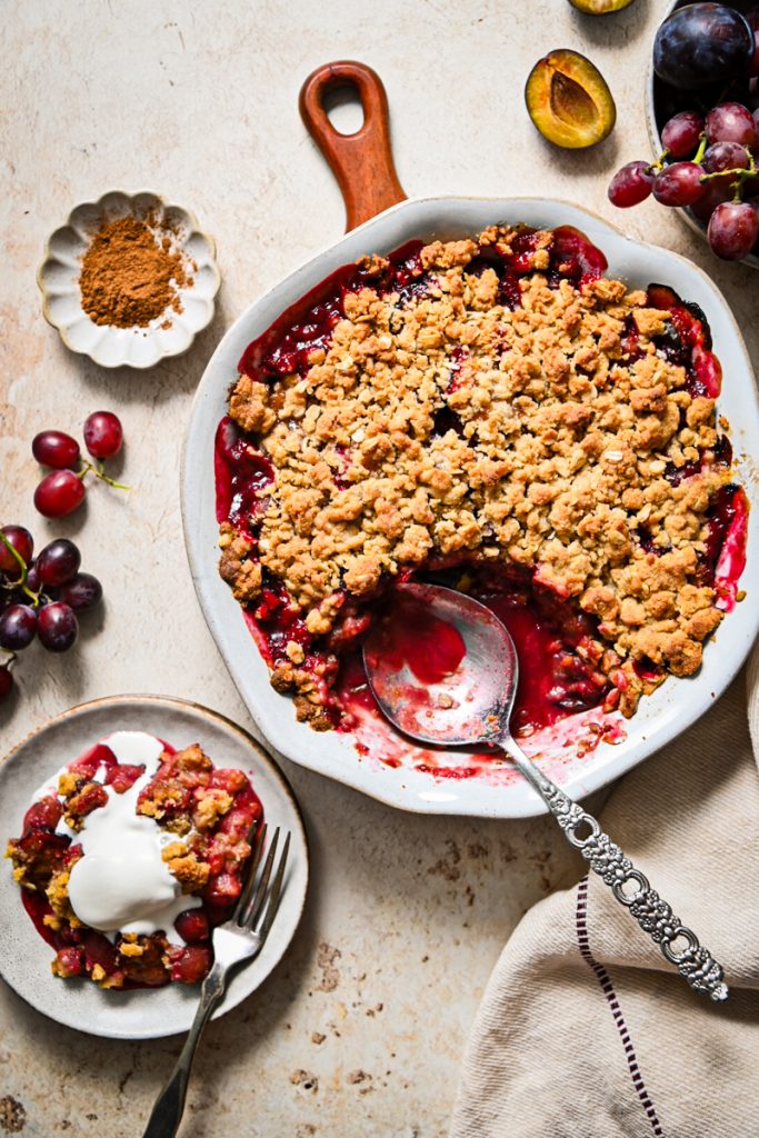 Overhead shot of plum and grape crisp in a pie dish with a portion missing, next to a plate with the portion.