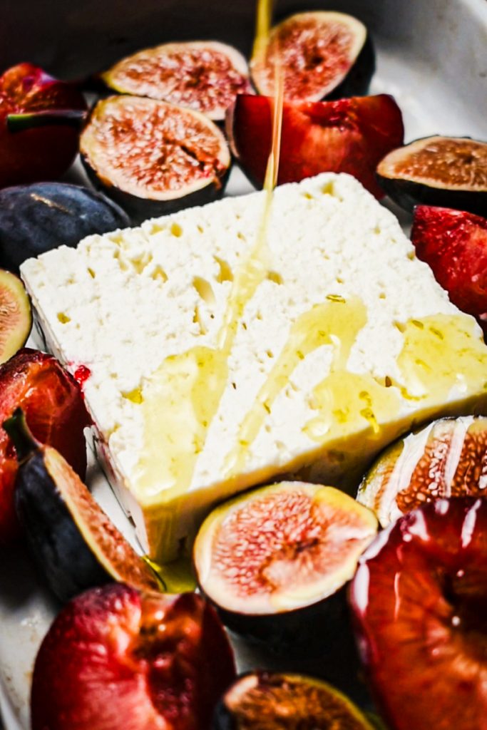Feta in a baking dish with figs and plums with olive oil being poured on top.