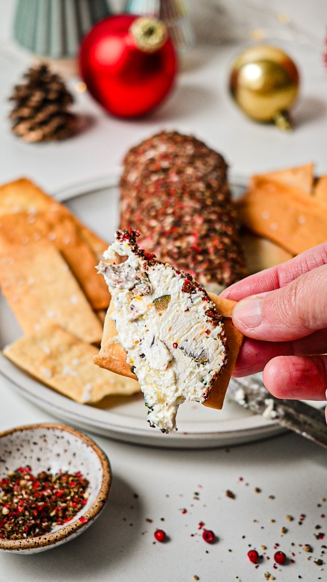 Hand holding a cracker with a slice of olive and feta cheese log on it.