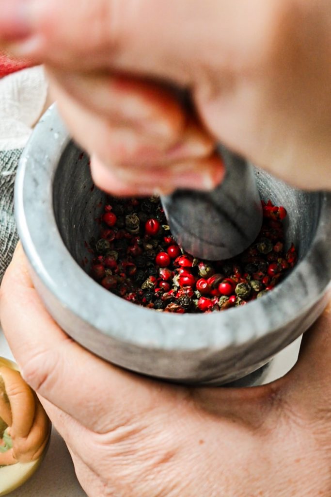 Hands using a mortar and pestle to crush a mixture of whole peppercorns.