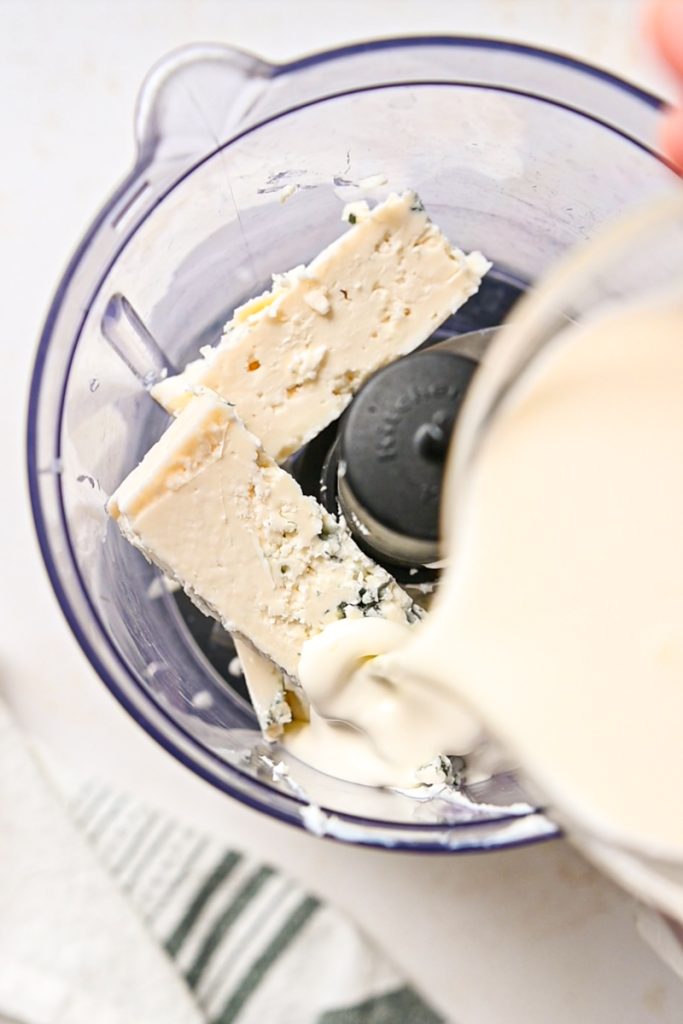 Pouring heavy cream into the gorgonzola cheese and cream cheese in a food processor.