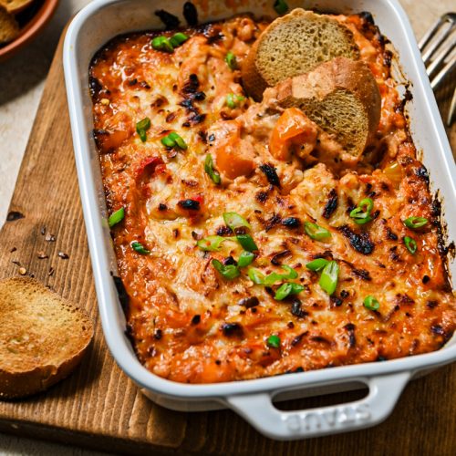 Baked ricotta dip with sweet peppers in a baking dish with two crostinis in the dip.