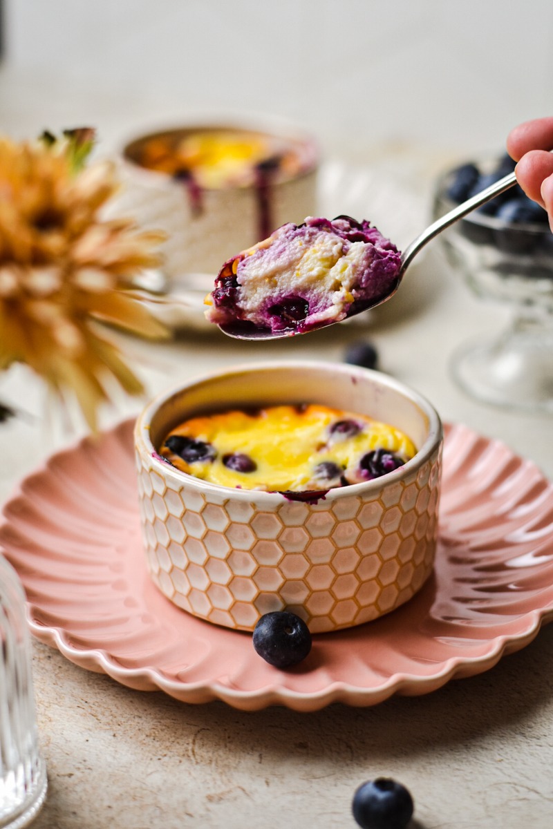 Spoonful of blueberry baked yogurt hovering over a ramekin of baked yogurt.