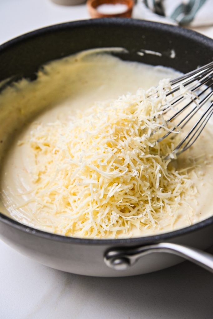 Bechamel sauce in a skillet with shredded cheese being added.