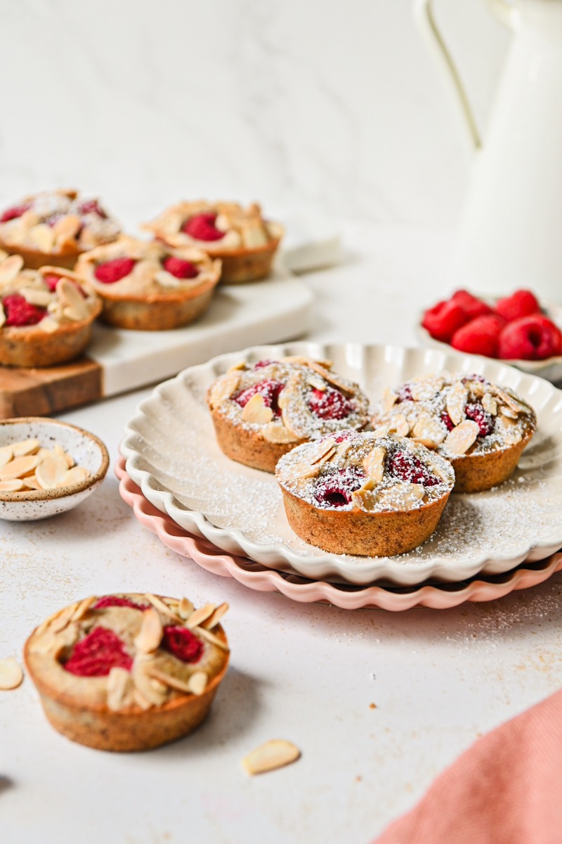 Finished raspberry financier muffins with some on a plate with powdered sugar on top.