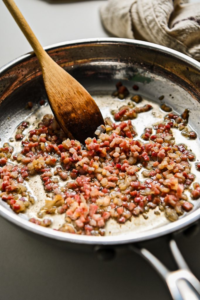 Cooking pancetta in a skillet with a wooden spoon.