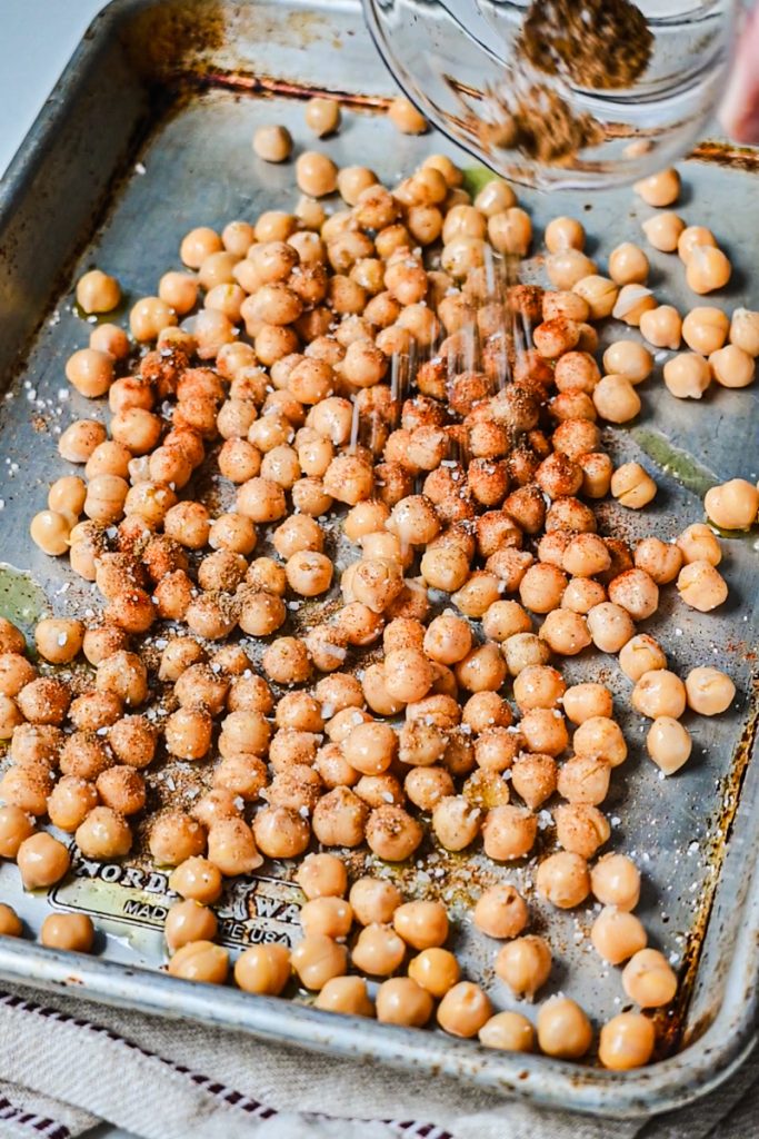 Sprinkling seasoning over the top of chickpeas on a sheet pan.