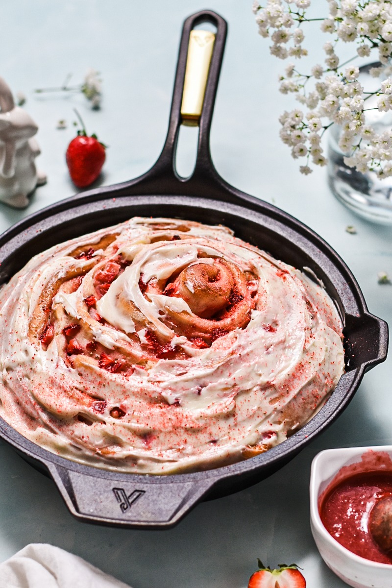 Side view of a giant strawberry roll cake in a cast iron skillet with cream cheese glaze on top.