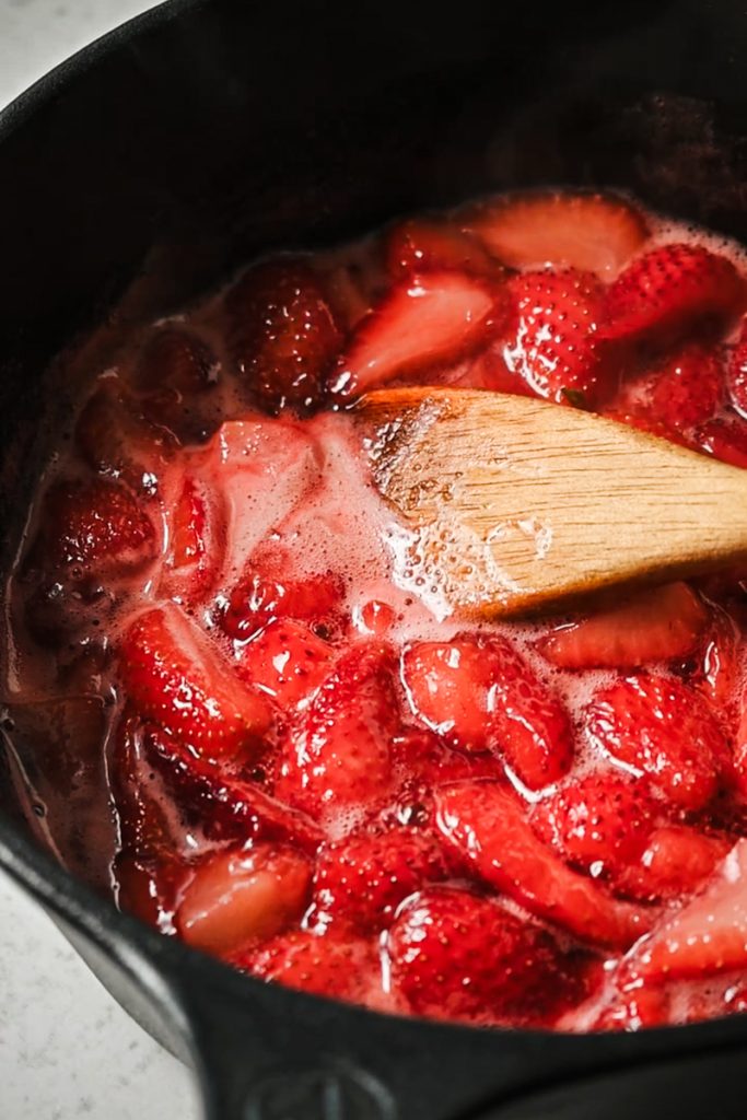 Strawberries simmering in a saucepan with a wooden spoon.