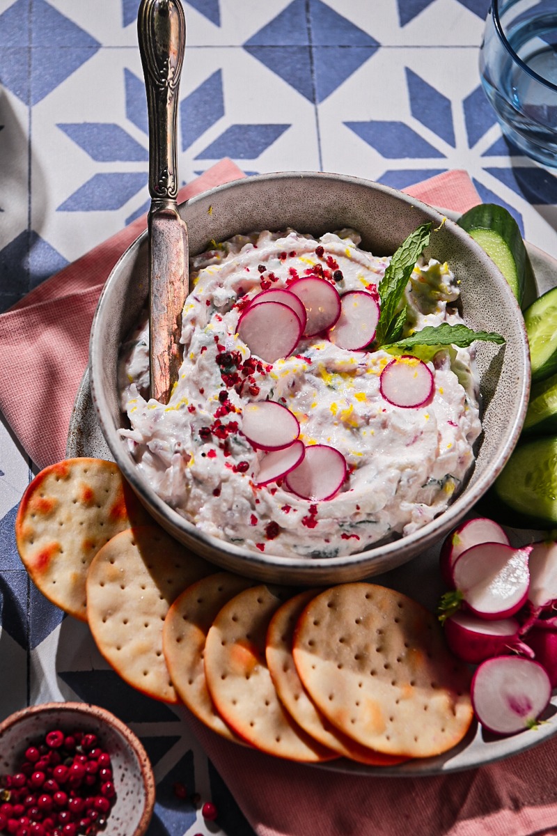 Side view of radish ricotta dip in a bowl garnished with radishes, herbs, and pink peppercorns.