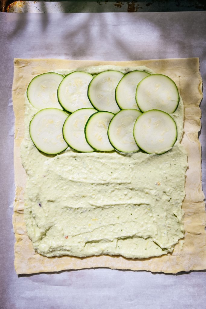 Puff pastry sheet with layer of pesto ricotta, topped with slices of zucchini.