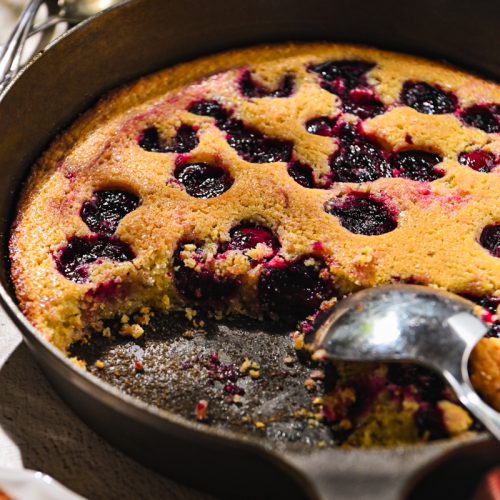 Serving spoon taking some cherry spoon cake from a cast iron skillet.
