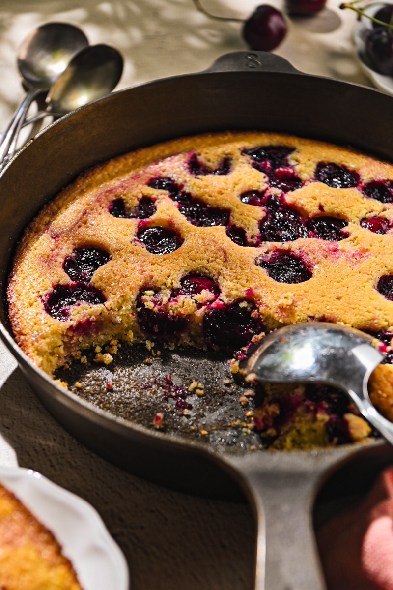 Serving spoon taking some cherry spoon cake from a cast iron skillet.