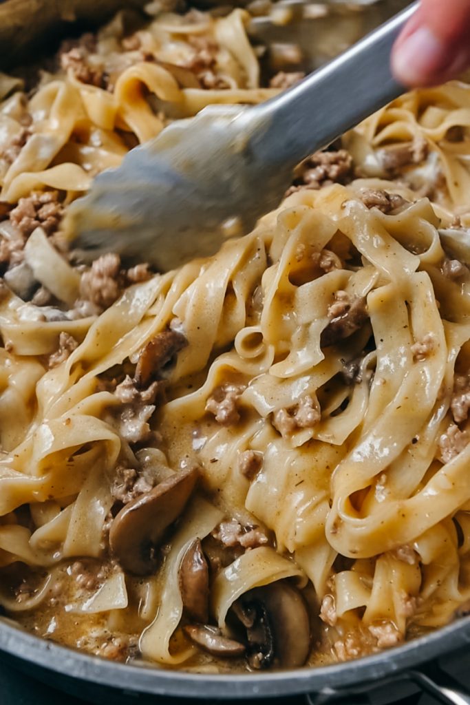 Using tongs to mix together the noodles and veal stroganoff in a skillet.