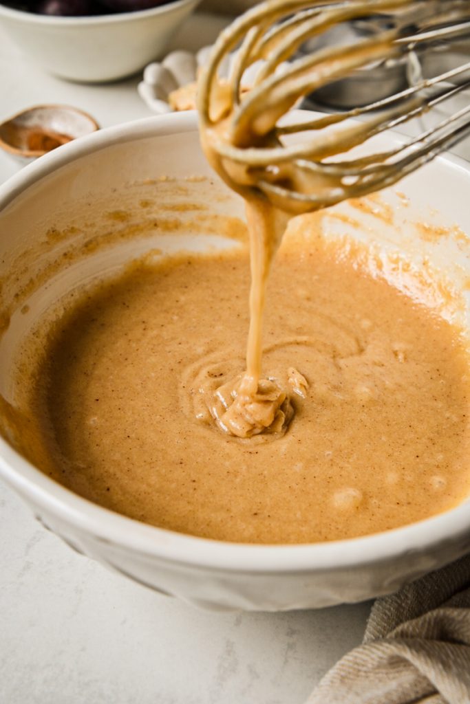 Whisk lifted out of the smooth almond cake batter in a white bowl.