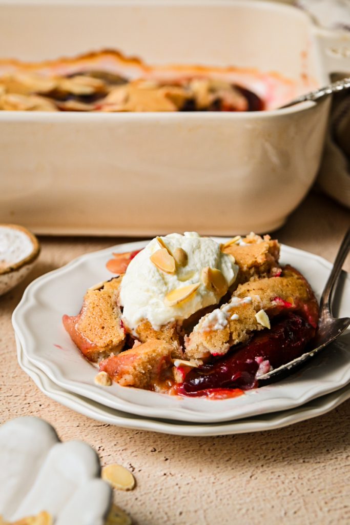 Slice of Plum Almond Cake topped with whipped cream and sliced almonds on a white plate.