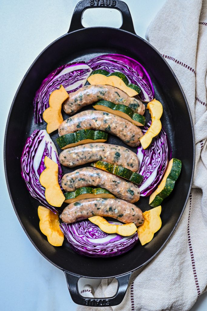Raw chicken sausages, acorn squash, and red cabbage wedges arranged on a sheet pan before roasting.